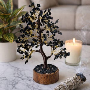 May include: A decorative tree sculpture with black gemstone leaves and gold wire branches. The tree sits in a wooden base filled with small black stones. A lit candle and a bundle of burning sage are also on the marble surface.