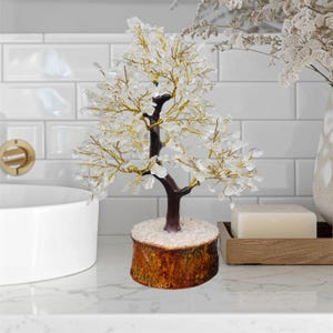 May include: A white crystal tree with gold wire branches and a brown wooden base. The tree is sitting on a white countertop.