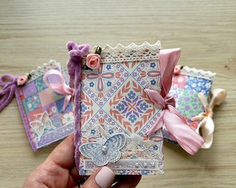May include: Three small rectangular notebooks featuring a repeating geometric tile pattern in shades of pink, blue, and white. Each notebook is decorated with a lace trim, a pink ribbon tie, a small fabric rose, and a butterfly cutout.