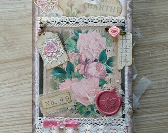 May include: A rectangular journal cover featuring a vintage floral rose print, layered with lace trim, a pink wax seal with a bee motif, a butterfly cutout, and a tag labeled No. 49. The cover includes a fabric ribbon pull and decorative paper elements.