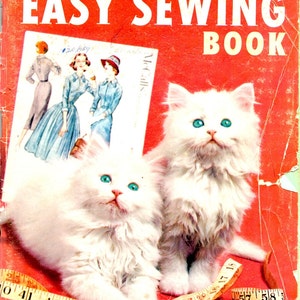 May include: Vintage McCall's Easy Sewing Book with two fluffy white kittens with bright blue eyes. The book cover is red with the title in white and a picture of women's fashion. A yellow measuring tape and spools of thread are in the foreground.