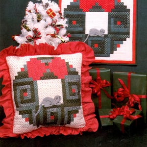 May include: A square pillow and a wall quilt featuring a holiday wreath pattern with a gray mouse applique. The designs use red, dark green, and white fabrics. The pillow has a ruffled red border. Text includes "HOLIDAY WREATH" and "Pattern C-15".