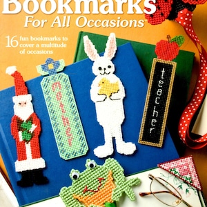May include: A collection of handmade plastic canvas bookmarks in various shapes, including a Santa, a frog, a bunny, and a ladybug. The bookmarks are in bright colors and are displayed on top of books. The text "Bookmarks For All Occasions" is visible.