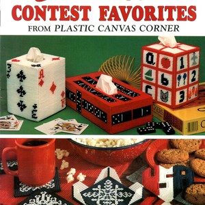 May include: A vintage craft book cover titled "Creative Contest Favorites" featuring plastic canvas projects. The image showcases tissue box covers, coasters, and other items with playing card, domino, and alphabet designs. A red mug and popcorn are also visible.