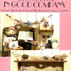 May include: A craft book cover with the title "Strings and Things II In Good Company" and a subtitle "33 Projects - Including Ideas for Garlands, Wreaths, Baskets, Centerpieces, and More". The cover features a photograph of a rustic setting with a wooden shelf, a small wooden wagon, and a variety of handmade items, including a heart-shaped pillow, a book, and a garland of fabric birds.