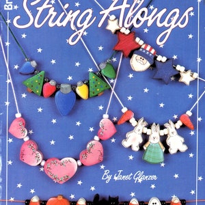 May include: A book cover titled "All Year String Alongs" featuring several handmade necklaces. The necklaces have various wooden charms, including Christmas trees, hearts, bunnies, and Halloween-themed shapes. The background is blue with white stars.