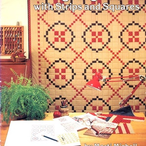 May include: A book titled "Designing Quilts with Strips and Squares" by Marti Michell. The cover features a close-up of a quilt with a geometric pattern of red, black, and white squares. The quilt is displayed on a table with a lamp, pencils, and a ruler.