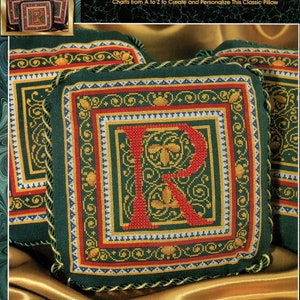 May include: Cross-stitch pillow with the letter "R" in red, surrounded by a green, gold, and red border. The pillow is square, with a decorative gold trim. The text "Elegant Alphabet" is at the top.
