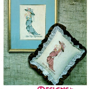 May include: A cross stitch pattern featuring a girl in a blue dress and a hat, with the text "Little Miss Make Believe". The pattern is framed and displayed on a brown burlap background.