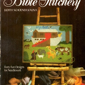 May include: A book titled "Bible Stitchery" by Judith Schoener Kalina. The cover features a colorful needlepoint design of Noah's Ark with animals, set against a brown background. The book title is in a decorative script font. The book is surrounded by colorful yarn skeins and a wooden easel.