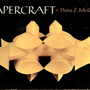 May include: A book cover with the title "PAPERCRAFT" and the author "Dona Z. Meilach". The cover features a geometric design of interlocking, golden-colored shapes against a black background.
