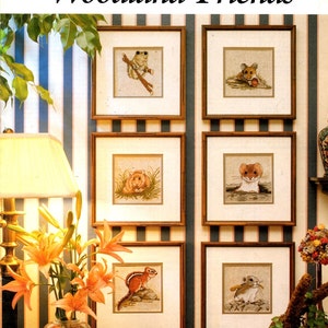 May include: A collection of framed cross-stitch artworks featuring woodland animals, including a frog, mouse, hamster, chipmunk, and other small creatures. The artwork is displayed on a striped wall, with the title "Woodland Friends" at the top.