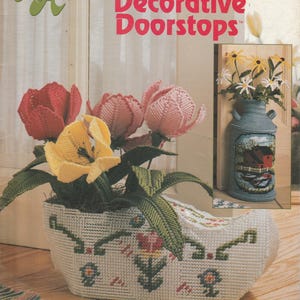 May include: A craft pattern cover featuring plastic canvas decorative doorstops. The cover displays a white, stitched planter with red, pink, and yellow flowers. A smaller image shows a blue milk can with flowers.