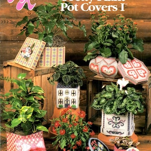 May include: A variety of plants in decorative plastic canvas pot covers. The covers feature various patterns, including plaid, floral, and heart designs. The image includes the text "Pretty Plant Pot Covers I" and the Annie's Attic logo.