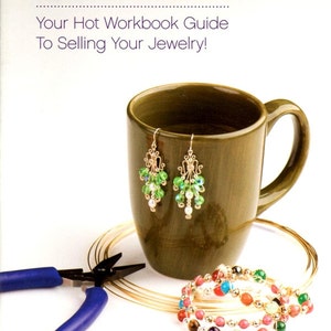 May include: A book cover titled Wire Java: Your Hot Workbook Guide To Selling Your Jewelry! by Mitzi McCartha. It features a brown ceramic mug, a pair of green and white beaded wire earrings, a coil of gold wire, and blue-handled pliers.