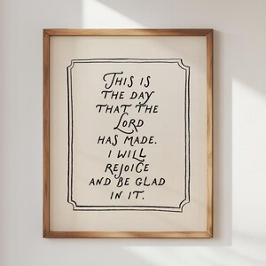 May include: A framed print with the text "THIS IS THE DAY THAT THE LORD HAS MADE. I WILL REJOICE AND BE GLAD IN IT." The text is in black script on an off-white background, framed in a light brown wooden frame.