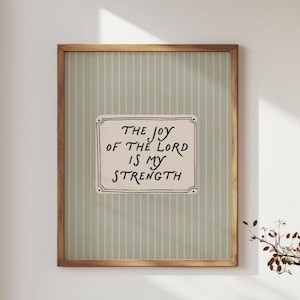 May include: A framed print with the text "THE JOY OF THE LORD IS MY STRENGTH." The artwork features a light green striped background and a cream-colored rectangle with black lettering. The frame is made of wood.