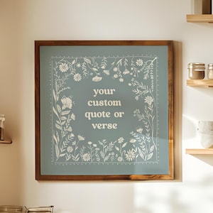 May include: A square wall art piece in a wooden frame featuring a muted sage green background with a white floral border and the centered text "your custom quote or verse" in a simple, clean, sans-serif font, displayed on a kitchen wall.