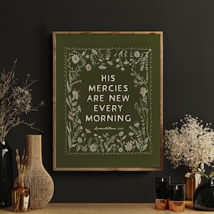 May include: A framed print with the text "HIS MERCIES ARE NEW EVERY MORNING" in white on a green background. The print is surrounded by a floral border. The image is displayed on a wooden cabinet with vases and candles.