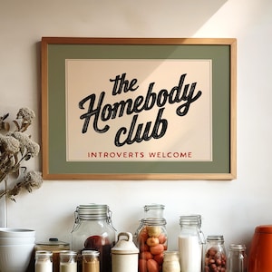 May include: A framed print with the text "the Homebody club" and "Introverts Welcome" hangs above a countertop. Various glass jars, an orange teapot, and a vase of dried flowers are arranged on the wooden surface.