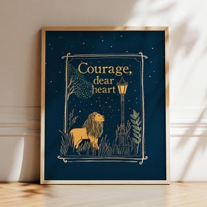 May include: A framed art print with a navy blue background. The artwork features a lion, a lamppost, and the words 'Courage, dear heart' in gold. The lion is depicted in a golden color, standing in a field of grass. The lamppost is black with a yellow light.
