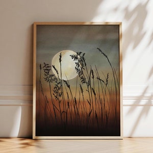 May include: A framed landscape art print featuring a large, full moon in a dusky sky. Silhouetted grasses and plants are in the foreground, with warm orange and brown tones at the bottom of the image. The artwork is in a light-colored wooden frame.