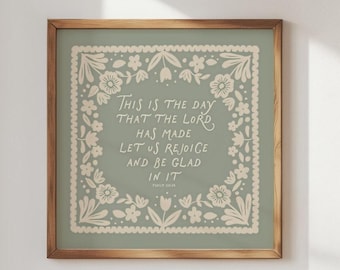 May include: A framed wall art piece with a sage green background and a floral border. The text "THIS IS THE DAY THAT THE LORD HAS MADE LET US REJOICE AND BE GLAD IN IT" is displayed in a white, elegant font. The frame is a warm, natural wood tone.