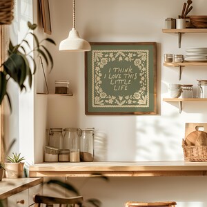 May include: A framed wall art piece with the text "I THINK I LOVE THIS LITTLE LIFE" in a white floral design on a green background. The art is displayed in a kitchen setting with wooden shelves, jars, and a woven basket.