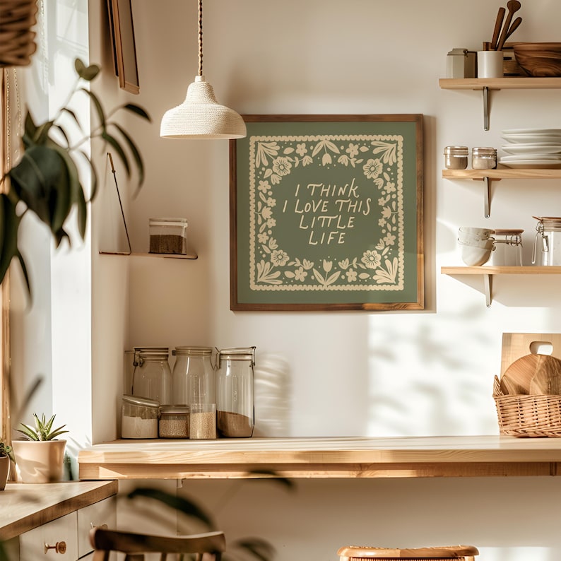 May include: A framed wall art piece with the text "I THINK I LOVE THIS LITTLE LIFE" in a white floral design on a green background. The art is displayed in a kitchen setting with wooden shelves, jars, and a woven basket.