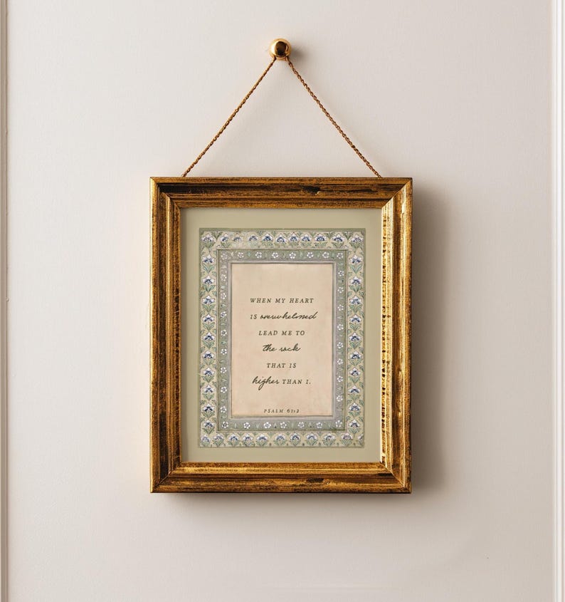 May include: A rectangular gold-toned frame hanging by a thin gold cord. Inside, a cream-colored print features a decorative floral border in shades of blue and green, surrounding the text: When my heart is overwhelmed lead me to the rock that is higher than I. Psalm 61:2.