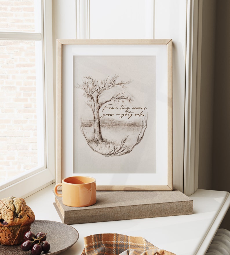 Puede incluir: Una impresi&oacute;n enmarcada con un dibujo a l&aacute;piz de un &aacute;rbol en un c&iacute;rculo con el texto "From tiny acorns grow mighty oaks". La impresi&oacute;n est&aacute; sobre un fondo blanco y est&aacute; enmarcada en un marco de madera clara. La impresi&oacute;n est&aacute; sentada en un alf&eacute;izar de ventana con una peque&ntilde;a taza naranja y un muffin.