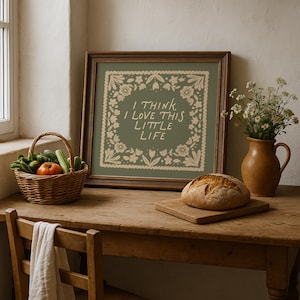 May include: A framed print with the text "I THINK I LOVE THIS LITTLE LIFE" surrounded by floral designs. A loaf of bread sits on a wooden cutting board. A basket of vegetables and a vase of flowers are also on the wooden table.