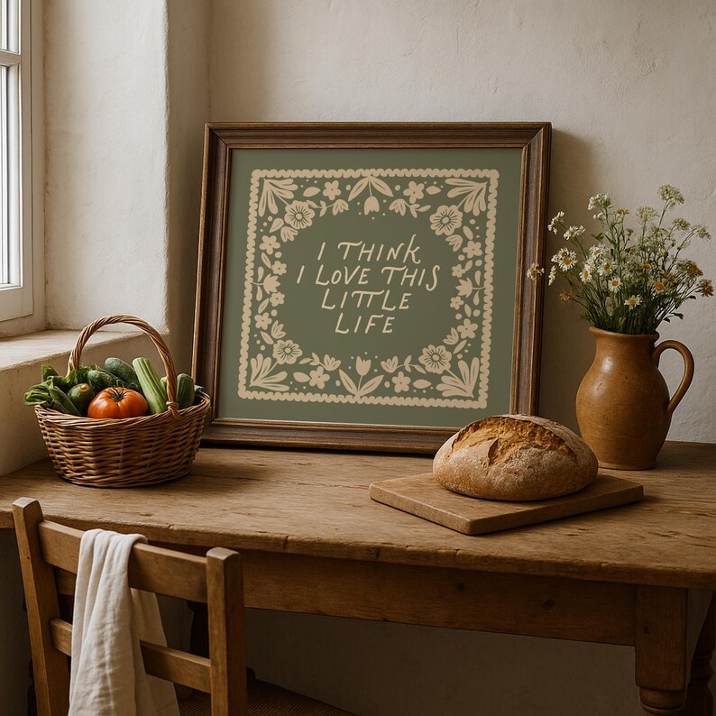 May include: A framed print with the text "I THINK I LOVE THIS LITTLE LIFE" surrounded by floral designs. A loaf of bread sits on a wooden cutting board. A basket of vegetables and a vase of flowers are also on the wooden table.