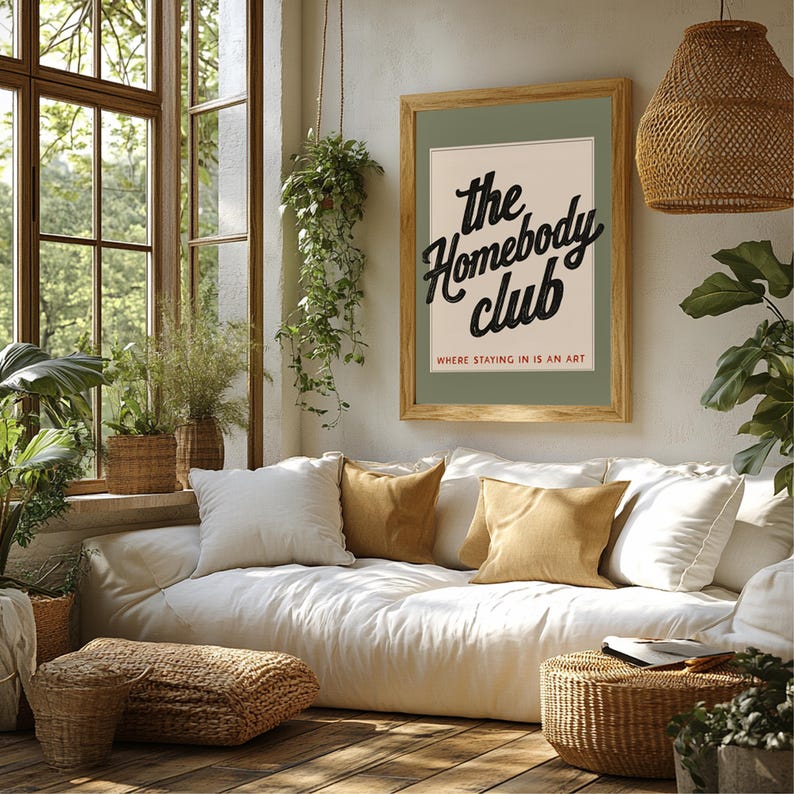K&ouml;nnte beinhalten: Ein gem&uuml;tliches Wohnzimmer mit gro&szlig;em Fenster, Pflanzen und einem gerahmten Poster. Das Poster zeigt "The Homebody Club" in schwarzer Schrift. Wei&szlig;e und ockerfarbene Kissen schm&uuml;cken ein wei&szlig;es Sofa. Geflochtene K&ouml;rbe und eine Rattanlampe.