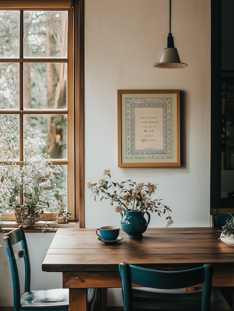 May include: A framed wall art print featuring the text When my heart is overwhelmed lead me to the rock that is higher than I Psalm 61:2, displayed in a light wood frame with a decorative border, hanging on a white wall above a wooden table.