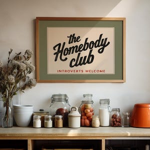 May include: A framed print with the text "the Homebody club" and "Introverts Welcome" in a kitchen setting. Various glass jars, a white ceramic bowl set, and an orange teapot are arranged on a wooden countertop. Dried flowers are in a clear vase.