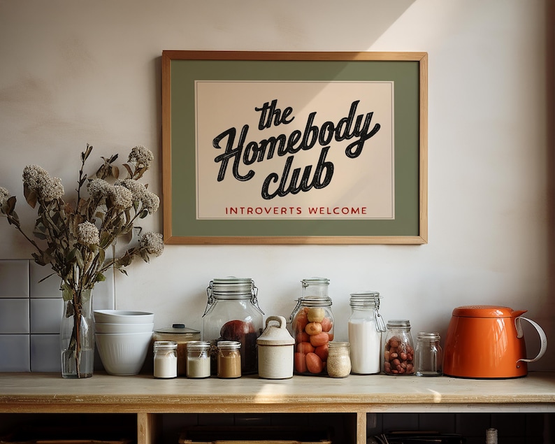 May include: A framed print with the text "the Homebody club" and "Introverts Welcome" in a kitchen setting. Various glass jars, a white ceramic bowl set, and an orange teapot are arranged on a wooden countertop. Dried flowers are in a clear vase.