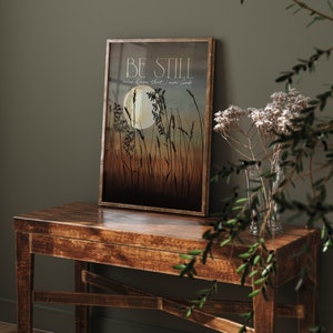 May include: A framed print with a quote that reads "Be still and know that I am God." The print features a silhouette of tall grass with a full moon in the background. The colors are muted and earthy, with shades of brown, blue, and white.
