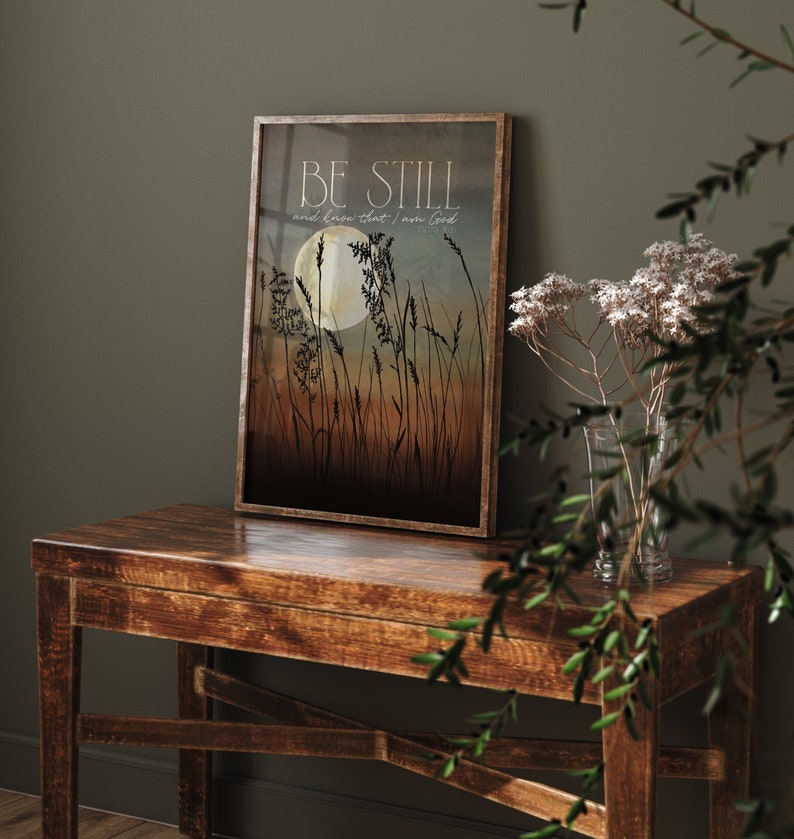 May include: A framed print with a quote that reads "Be still and know that I am God." The print features a silhouette of tall grass with a full moon in the background. The colors are muted and earthy, with shades of brown, blue, and white.