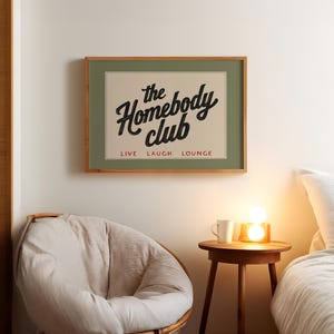 May include: A framed print with the words "the Homebody club" in a vintage style font. The print is in a wooden frame and hangs above a round chair and a small wooden table with a lamp and a mug.