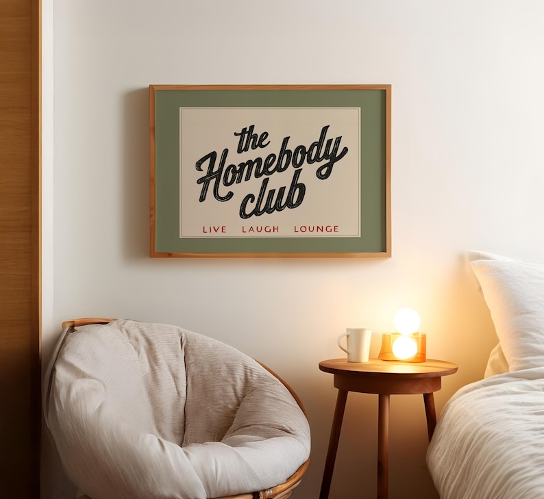 May include: A framed print with the words "the Homebody club" in a vintage style font. The print is in a wooden frame and hangs above a round chair and a small wooden table with a lamp and a mug.