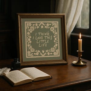 May include: A framed print with the text "I THINK I LOVE THIS LITTLE LIFE" surrounded by floral designs. The print is on a wooden table with an open book, inkwell, and a lit candle in a brass holder.