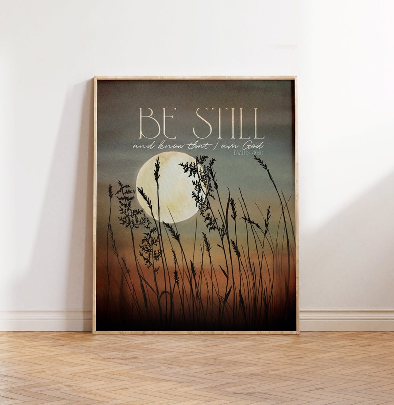 May include: A beige and brown watercolor print with a full moon and tall grass silhouetted against a sunset sky. The print features the text "Be Still and know that I am God" with the scripture reference "Psalm 46:10".