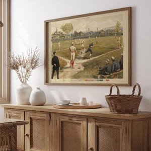 May include: A framed vintage baseball print depicting a game in progress. The artwork features players in period uniforms on a green field, with spectators and a wooden frame. The color palette includes greens, browns, and whites.
