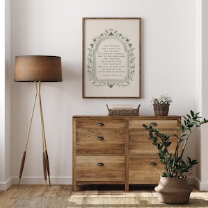 May include: A framed print with a poem in green lettering, displayed above a wooden dresser. The poem is surrounded by a decorative green border. A floor lamp with a brown shade and a potted plant are also visible.