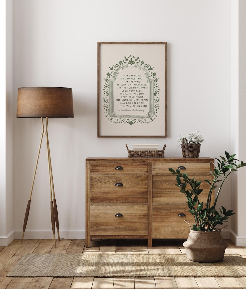 May include: A framed print with a poem in green lettering, displayed above a wooden dresser. The poem is surrounded by a decorative green border. A floor lamp with a brown shade and a potted plant are also visible.