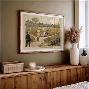 May include: A framed vintage baseball print depicting a game in progress. The artwork features players in period uniforms on a green field, with spectators and a wooden frame. The print is displayed on a wooden surface.