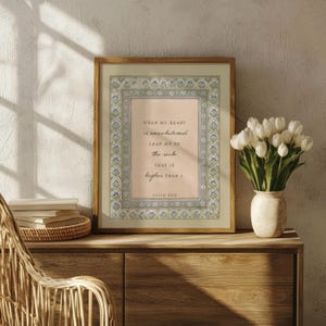 May include: A framed wall art print featuring a light pink background with the text When my heart is overwhelmed lead me to the rock that is higher than I Psalm 61:2, surrounded by a decorative border of blue and green floral patterns.