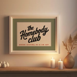 May include: A framed print with the text "the Homebody Club" in a black script font, with the tagline "WHERE STAYING IN IS AN ART" in a serif font. The print is displayed above a wooden surface with candles and a vase.