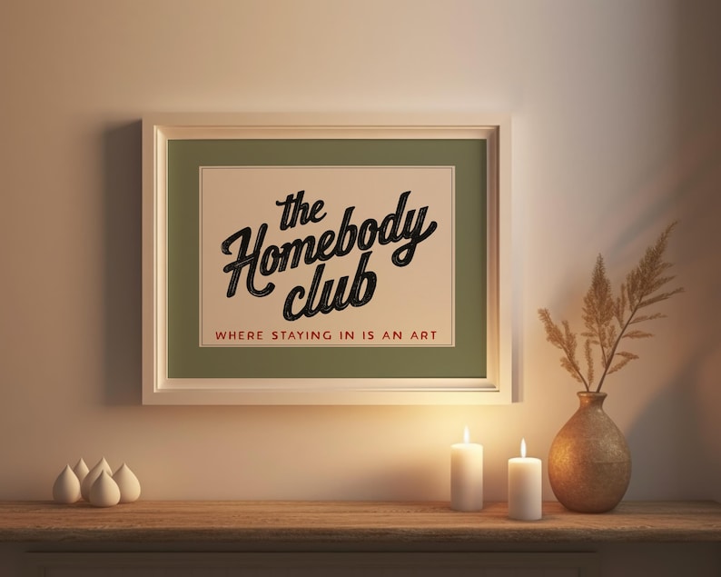 May include: A framed print with the text "the Homebody Club" in a black script font, with the tagline "WHERE STAYING IN IS AN ART" in a serif font. The print is displayed above a wooden surface with candles and a vase.